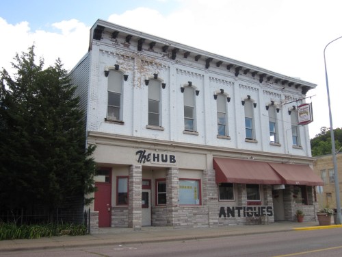 A historic building with a white facade, featuring "The Hub" and "Antiques" signs, surrounded by trees and blue sky.