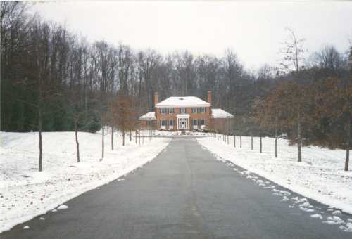 A large brick house at the end of a snowy driveway, surrounded by bare trees and a winter landscape.