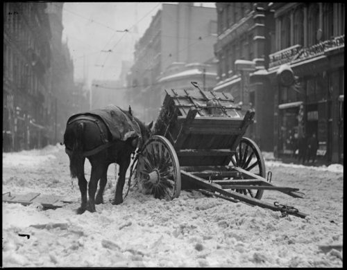 A horse stands beside a snow-covered cart on a deserted street, surrounded by buildings in a winter scene.