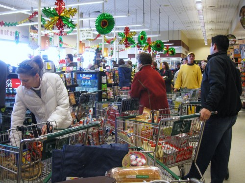 A busy grocery store with shoppers and carts, decorated for the holidays, creating a festive atmosphere.