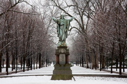 A snow-covered park with a statue of a figure with outstretched arms, surrounded by bare trees.