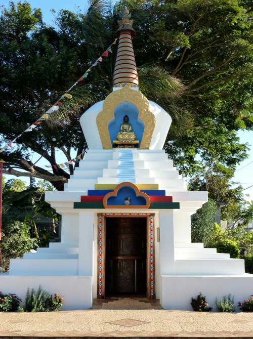 A colorful stupa with a golden Buddha statue at the top, surrounded by greenery and decorative flags.