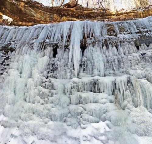 A frozen waterfall cascading down a rocky cliff, surrounded by icy formations and bare trees above.