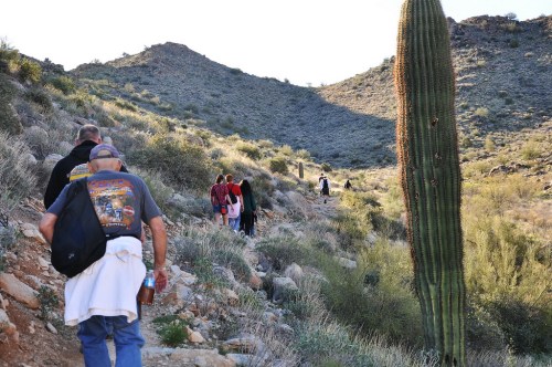A group of hikers walking along a rocky trail in a desert landscape with cacti and hills in the background.