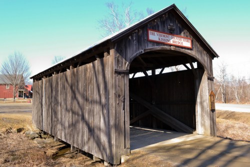 A rustic wooden covered bridge with a sign reading "Kissing Bridge" against a clear blue sky.