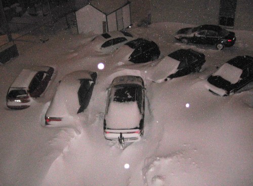 A parking lot filled with cars buried under heavy snow, with only a few visible above the snowdrifts.