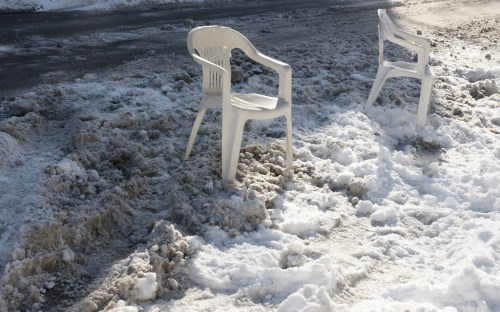 Two white plastic chairs sit on a snowy, slushy surface beside a road.
