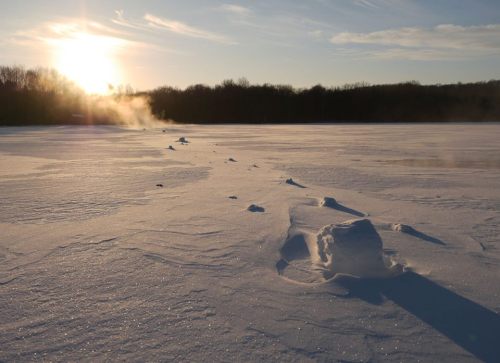 A snowy landscape at sunrise, with mist rising from a frozen lake and trees in the background.