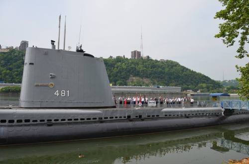 A submarine docked by a river, with a group of people in formal attire standing on the pier nearby.
