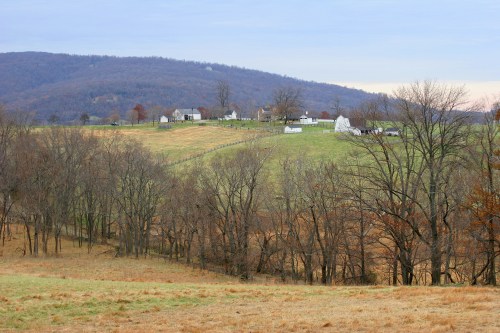 A serene landscape featuring rolling hills, bare trees, and a distant farmstead under a cloudy sky.