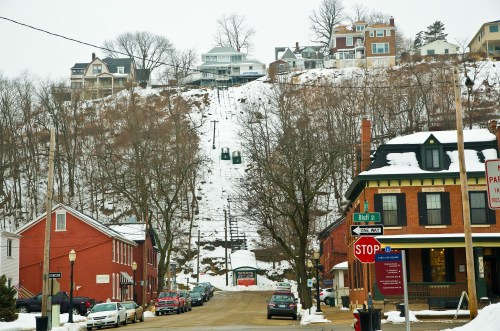 A snowy street scene with houses on a hill, a funicular railway, and parked cars in a small town.