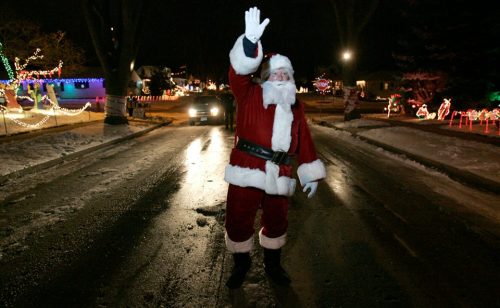 Santa Claus waves while standing in a snowy street decorated with colorful Christmas lights at night.