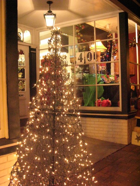 A lit wire Christmas tree stands outside a shop, with festive decorations visible through the window behind it.
