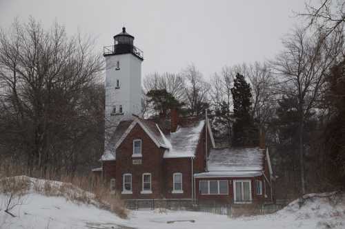 A snowy scene featuring a tall lighthouse beside a brick house, surrounded by bare trees and a winter landscape.