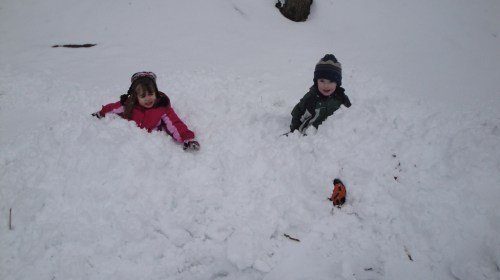 Two children play in deep snow, one in a pink jacket and the other in a green coat, with a toy nearby.