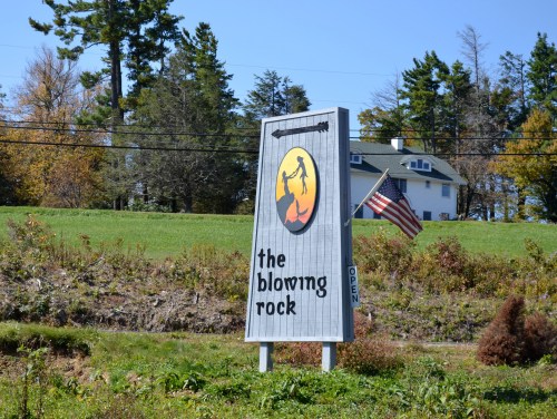 Sign for "The Blowing Rock" with an American flag, set against a green landscape and trees in the background.