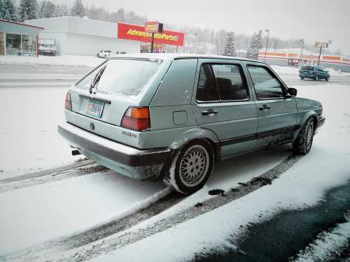 A light blue car parked on a snowy road, with tire tracks leading away in a wintery landscape.