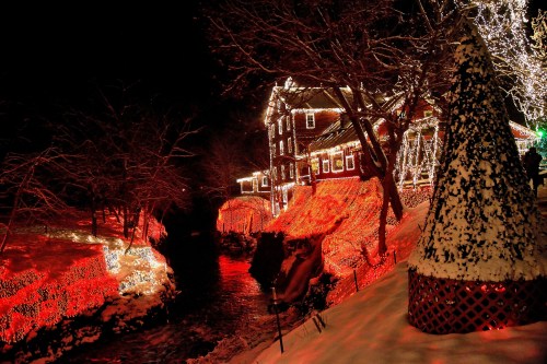A snowy scene at night featuring a house adorned with colorful Christmas lights and a glowing stream nearby.