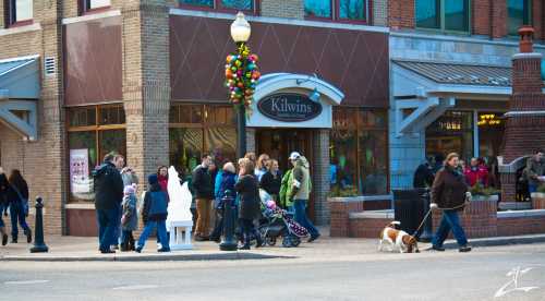 A busy street scene with people gathered outside Kilwins, a festive shop, and a dog walking by.