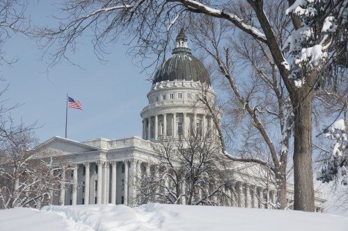 The Utah State Capitol building surrounded by snow, with a clear blue sky and an American flag flying.