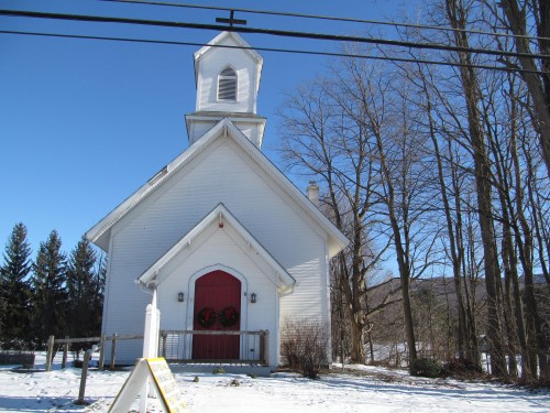 A white church with a red door and cross, surrounded by snow and trees under a clear blue sky.