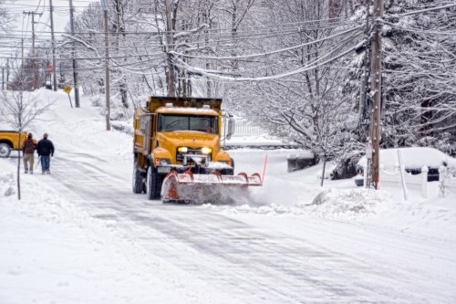 A snowplow clears a snowy street while a person walks nearby, surrounded by snow-covered trees and power lines.