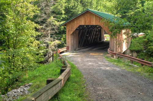A wooden covered bridge surrounded by lush greenery and a gravel path leading into it.