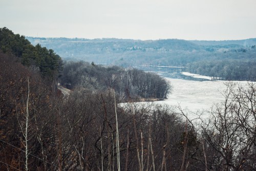 A serene winter landscape featuring a frozen river surrounded by bare trees and distant hills under a cloudy sky.