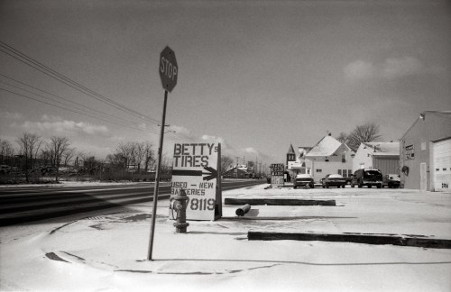 Black and white photo of a snowy street with a "Betty Tires" sign and a stop sign, featuring nearby buildings.