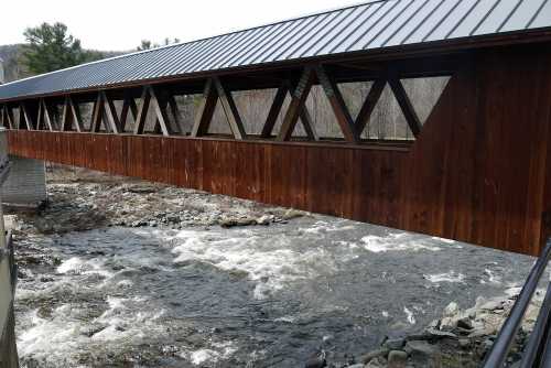 A wooden covered bridge spans a rushing river, surrounded by trees and a clear sky.