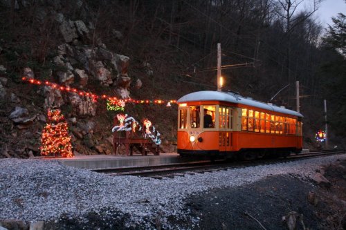 A vintage orange trolley car stops at a festive station adorned with holiday lights and decorations.