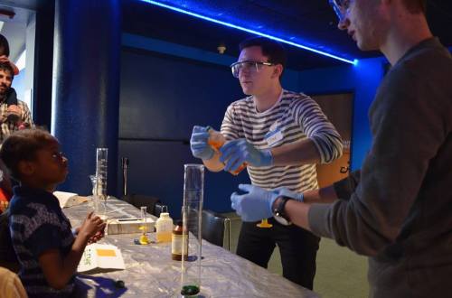 A child watches as two adults conduct a science experiment with colorful liquids at a table.