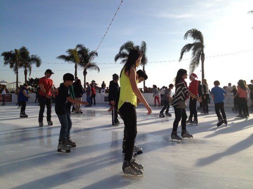 A lively outdoor ice skating rink with people of all ages skating under a clear sky and palm trees.