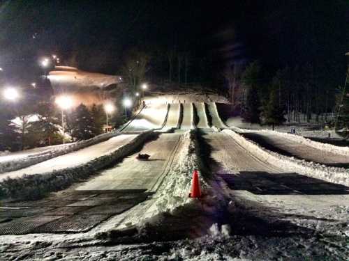 Snow tubing hill at night, featuring multiple lanes and illuminated by lights, surrounded by trees.