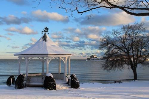 A snow-covered gazebo by a calm lake, with a tree and clouds in the background on a winter day.