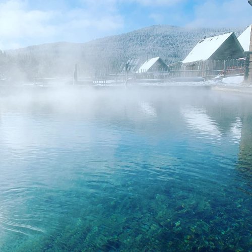 A serene hot spring surrounded by snow-covered mountains and mist, with wooden structures in the background.