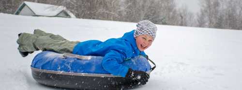 A child in a blue jacket joyfully sleds down a snowy hill on a tube, surrounded by falling snowflakes.