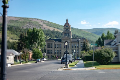 Historic building with a clock tower, surrounded by trees and mountains, along a quiet street on a sunny day.