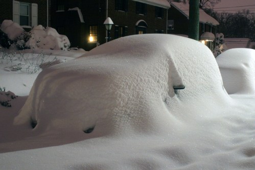 A car completely covered in thick snow, with only its outline visible, in a residential area at night.
