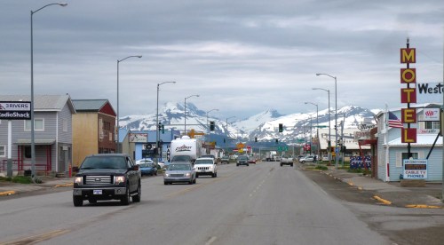 A small town street with shops and a motel, set against a backdrop of snow-capped mountains under a cloudy sky.