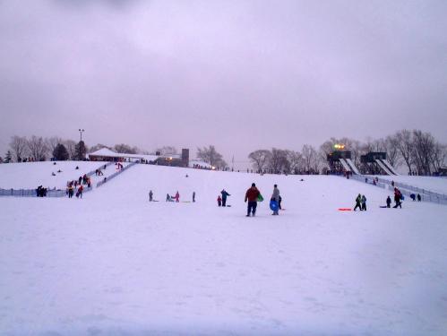 A snowy hill filled with people sledding and walking, with trees and a ski lift in the background under a cloudy sky.
