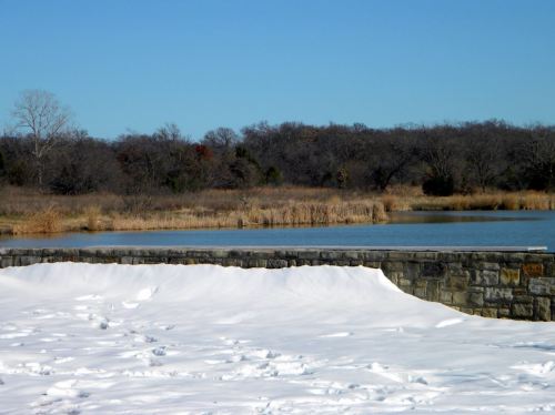 A serene landscape featuring a snow-covered foreground, a stone wall, and a calm lake surrounded by bare trees.