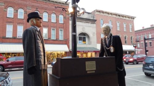 Two life-sized figures in historical attire stand near a lamppost on a street, with shops visible in the background.