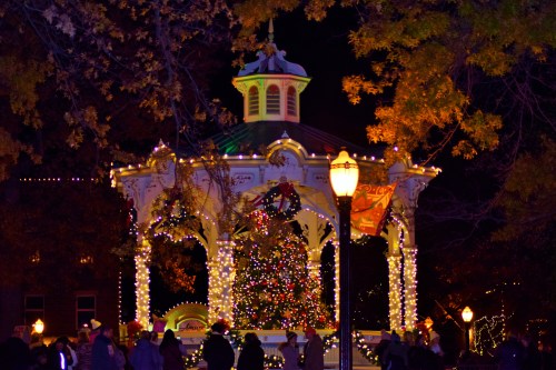 A beautifully lit gazebo adorned with holiday decorations and a Christmas tree, surrounded by people at night.