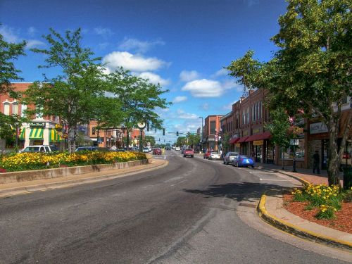 A vibrant town street lined with shops, flowers, and trees under a blue sky with fluffy clouds.