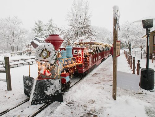 A festive train decorated with lights and a wreath, parked in a snowy landscape with trees and a sign nearby.