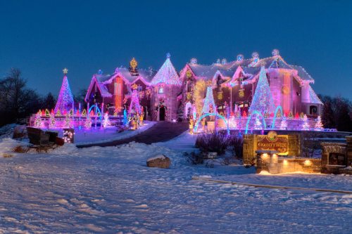 A brightly lit house decorated with colorful Christmas lights, surrounded by snow and festive decorations.