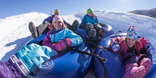 Four children and an adult enjoy tubing down a snowy hill on a sunny day, smiling and wearing colorful winter gear.