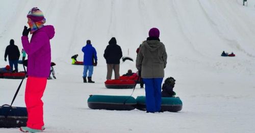 People in winter clothing enjoy snow tubing on a snowy hill, with some waiting and others sliding down.