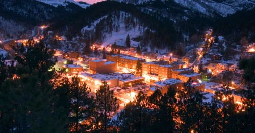 Aerial view of a snowy town at dusk, illuminated by warm lights, surrounded by mountains and trees.
